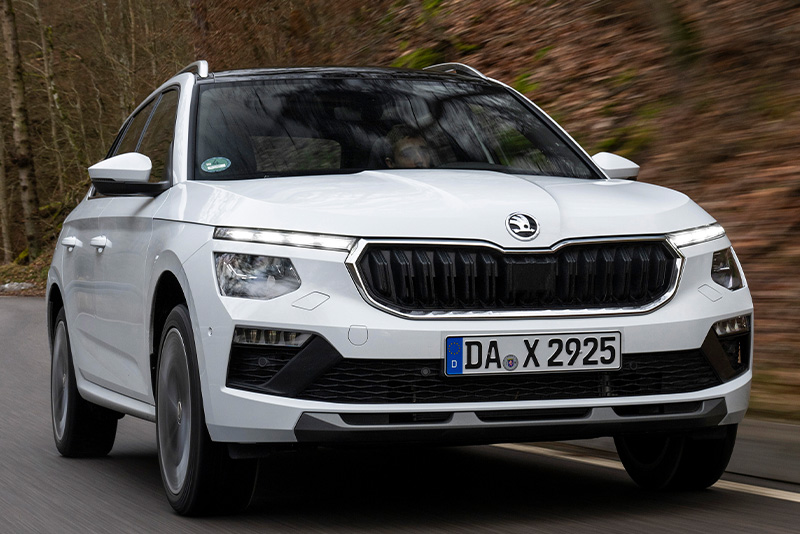 Wei&szlig;er Skoda Kamiq in der Dreiviertelansicht von vorne beim Fahren auf einer Landstra&szlig;e mit B&auml;umen im Hintergrund