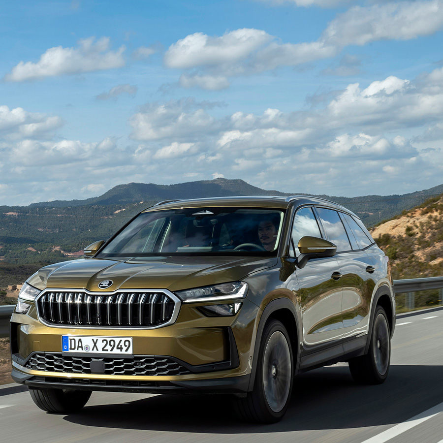 Ein gr&uuml;ner Skoda Kodiaq in der Dreiviertelansicht von vorne beim Fahren auf einer Landsta&szlig;e mit hohem Tempo mit einer bergigen Landschaft im Hintergrund