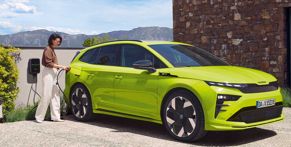 Seitenansicht eines Enyaq Coupe RS in gr&uuml;n metallic neben einer Elektrolades&auml;ule mit einer Frau daneben