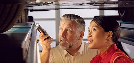 Ein Werkstattmitarbeiter zeigt einer Kundin mit einer Taschenlampe bei einem sich auf der Hebeb&uuml;hne befindenden Auto etwas am Boden des Autos